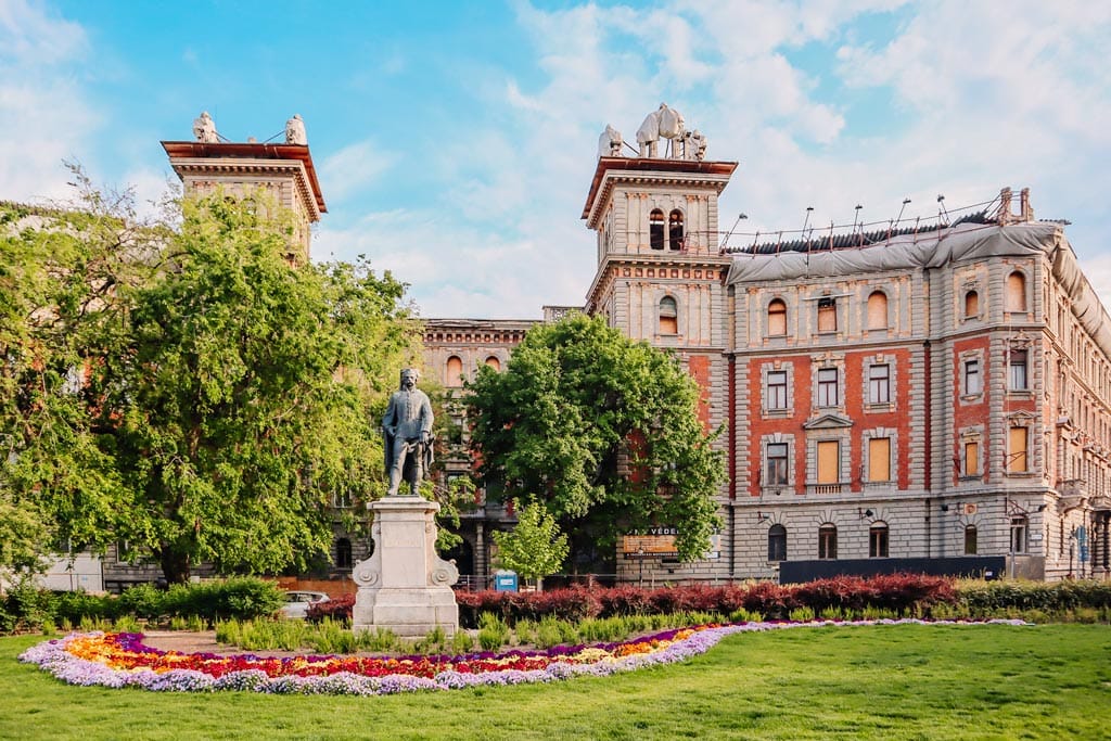 A square in Budpest with statue surrnounded by flowers and a beautiful building behind